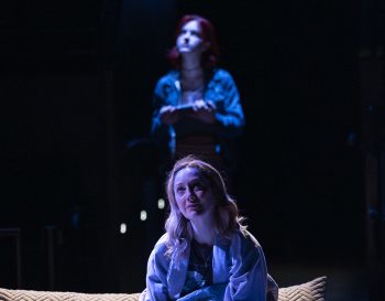 A woman sits in a bed while a younger girl looms behind her in the shadows.