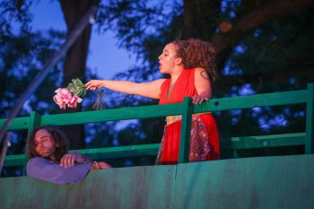 An actor on an elevated platform holds a bouquet of pink roses over another actor's head, who is hanging on to the bottom of the platform.