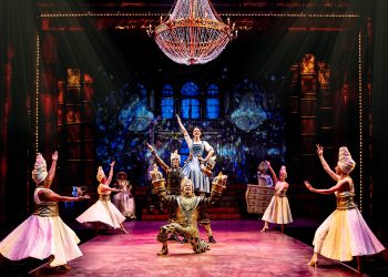 Belle and Lumiere dance in the middle of Beast's castle under a chandelier with dancers surrounding them.
