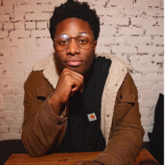 Devon Walker wearing glasses with his hand on his chin, sitting in front of a brick wall.