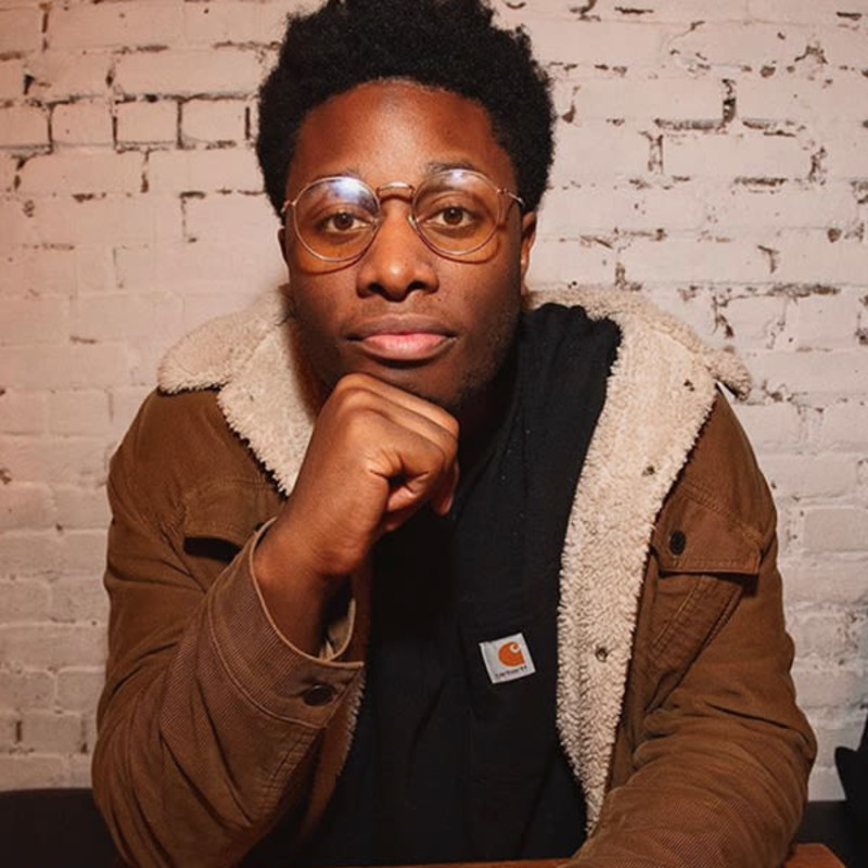 Devon Walker wearing glasses with his hand on his chin, sitting in front of a brick wall.