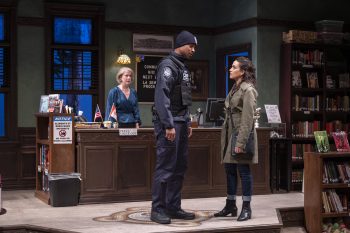 A woman and security guard look at each other in a library while a librarian looks at both from behind a check out desk.
