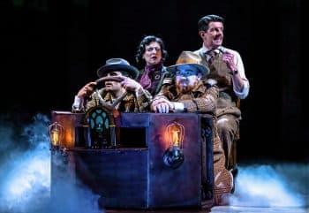Two men sit in front of a theater's set train car with hands on the steering wheel while behind a man and woman look around in suspense.