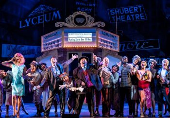 A cast of characters in dressy clothes on stage in front of a theater marquee