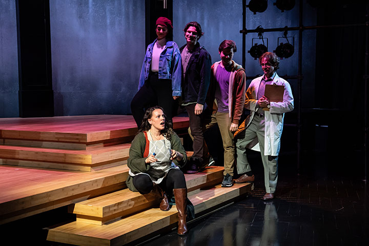 A woman sings on a stage seated on a step while a row of people stand behind her watching.