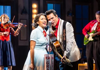 Two actors sing together into a microphone while behind them a violinist and guitarist play.