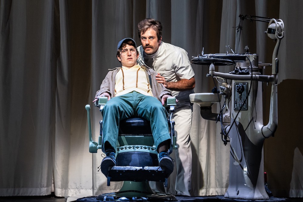 A patient in an operating chair next to a doctor standing next to medical equipment.