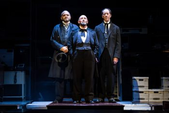 Three men in old-fashioned suits stand looking forward on a dark stage.