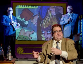 A man discusses a zoomed in comic panel, while two men in suits observe him.