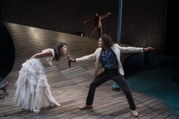 A man holds a mic up to a woman who is bending forward, in front of a sloped stage with a man balancing on it.
