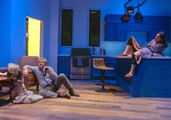 A man sits on a wooden floor drinking a bottle of beer while a woman watches while laying back on a kitchen counter