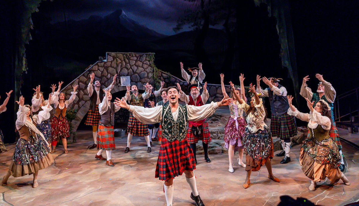 The cast of Brigadoon wearing Scottish garb and holding their hands up together.