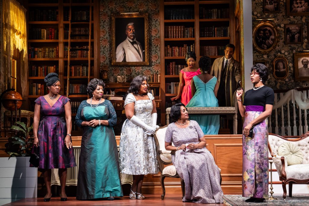 Women dressed in elegant clothing stand around a fancy parlor. 