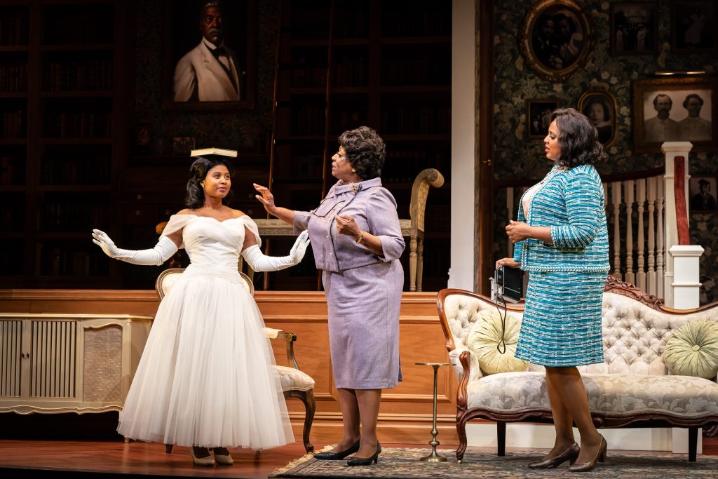 A woman in a fancy white dress balances a book on her head while two older women observe.