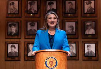 A woman in a blue suit jacket stands at a podium in front of a wall of war heroes.