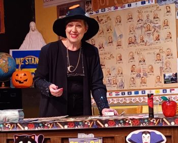 A woman dressed in black as a witch stands at the front of a classroom filled with Halloween decor.