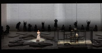 A woman in the spotlight on a stage with people climbing walls behind her and a room of cages next to her.