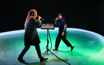 Two women on a green stage walk in circles around a piece of audio machinery on a stand.