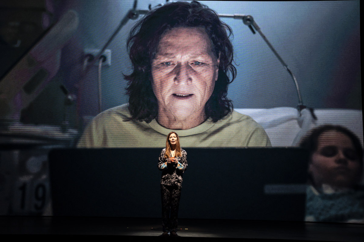 An actor stands in the middle of a dark stage with a large background photo showing a woman on a computer.