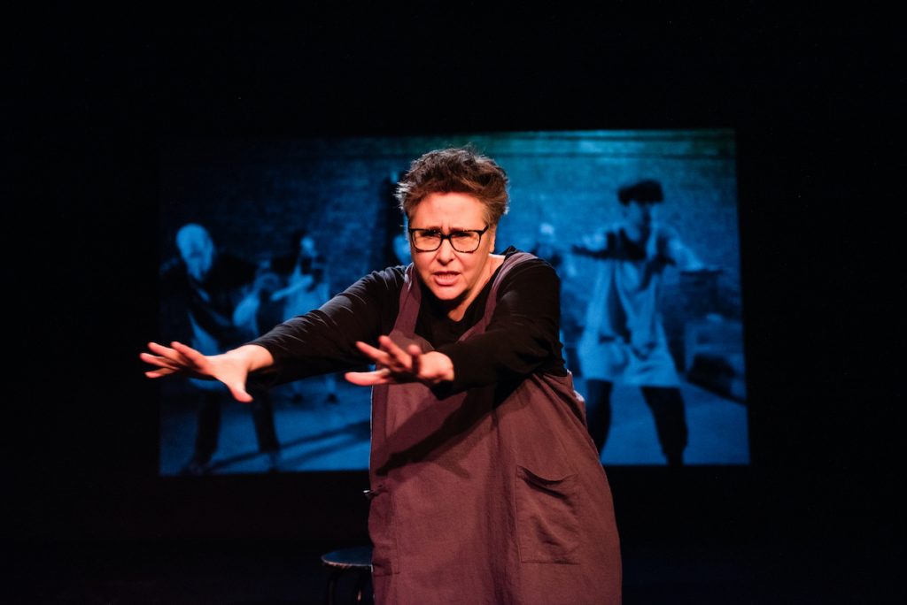 An actor reaches out towards the audience, with a projection of people dancing behind them.