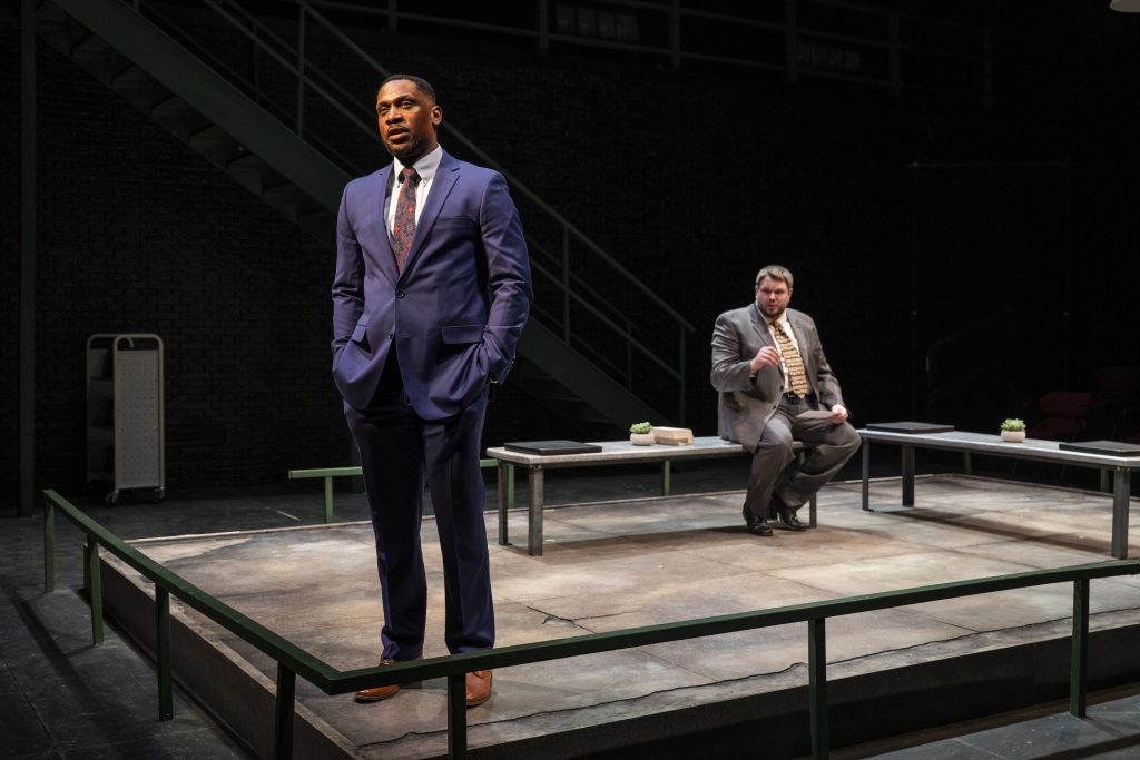 A man in a suit stands in the foreground looking out while another man sits behind him.