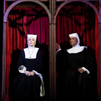 Two women dressed as nuns sit in a wooden confessional booth with red curtains, one looking serious and the other smiling and gesturing. Shadows of crosses are visible on the backdrop. Stage lighting highlights their faces.