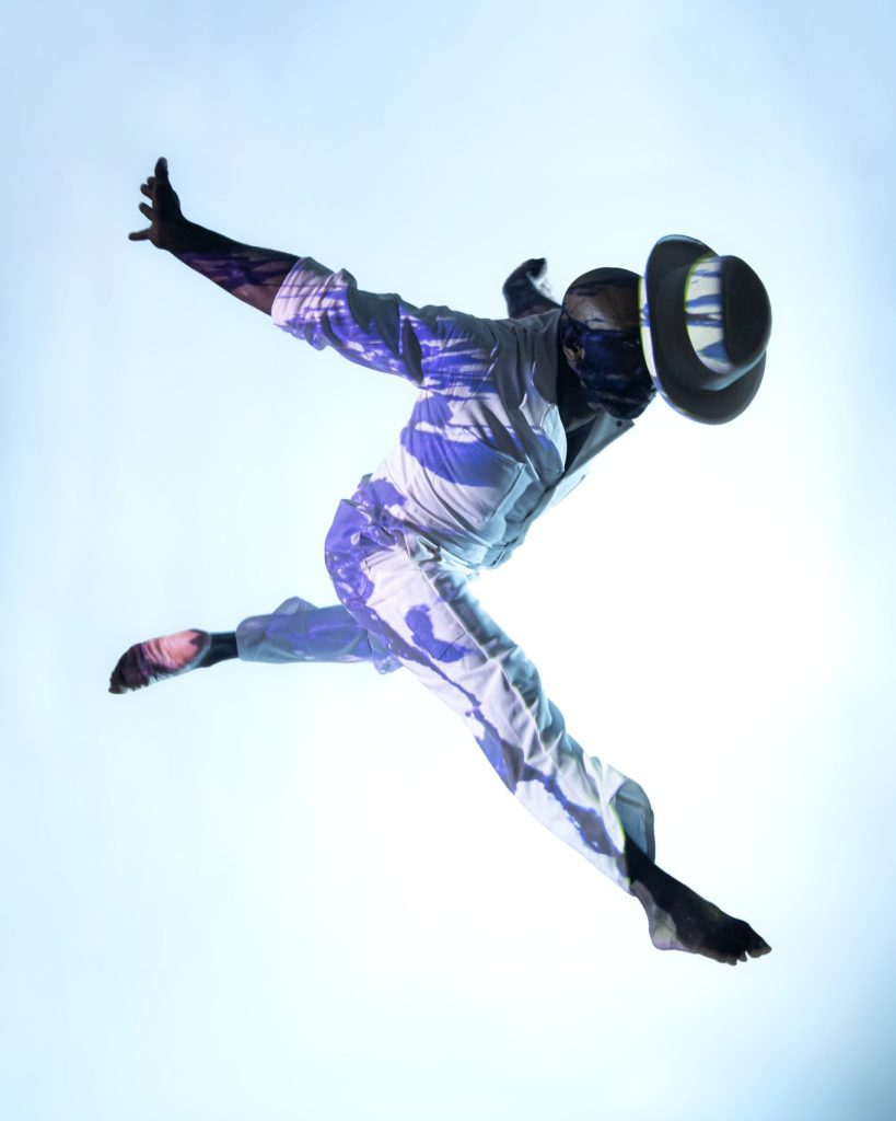A person in a hat and white clothing leaps in the air with arms and legs extended, creating a dynamic, diagonal pose against a bright, gradient background. Their face is obscured by shadows and the brim of the hat.