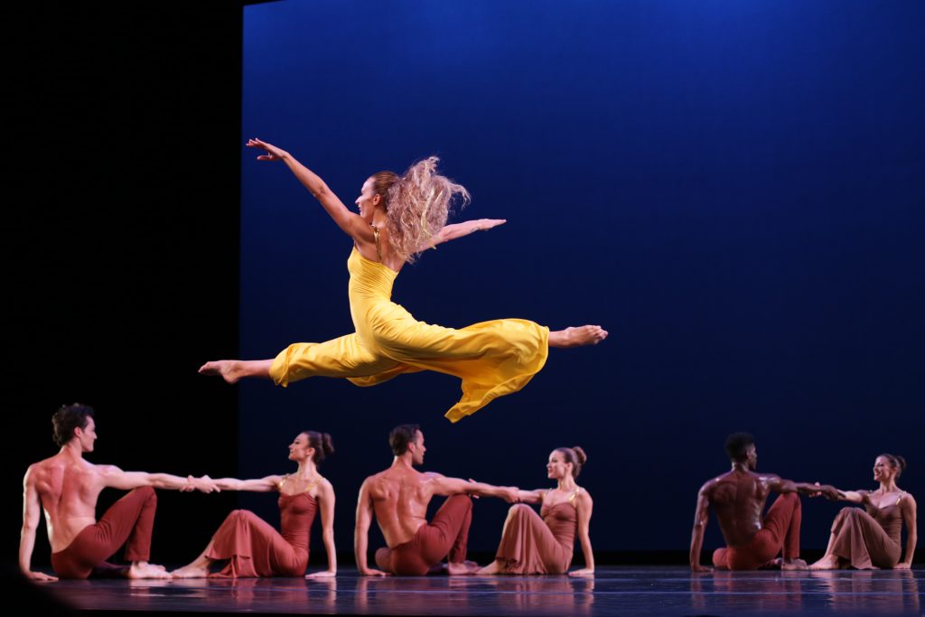 A female dancer in a yellow dress leaps gracefully mid-air on stage, surrounded by six seated dancers in earth-toned costumes, holding hands against a deep blue backdrop.