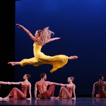 A female dancer in a yellow dress leaps gracefully mid-air on stage, surrounded by six seated dancers in earth-toned costumes, holding hands against a deep blue backdrop.