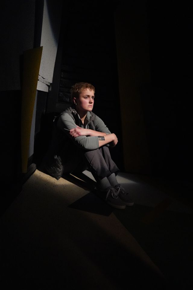 A person with short hair sits on the floor in a dimly lit room, hugging their knees to their chest and looking thoughtfully into the distance. Shadows and light create a dramatic effect around them.