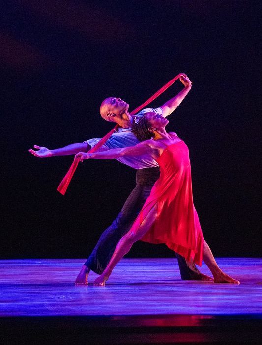 Two dancers perform on stage; one in a flowing red dress extends gracefully forward, while the other stands behind, arms wide, both holding a red scarf. The lighting creates a dramatic and artistic atmosphere.