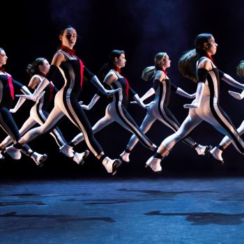 Seven dancers in matching black and white outfits and red neckties leap in unison on a stage, captured mid-air against a dark, smoky background.