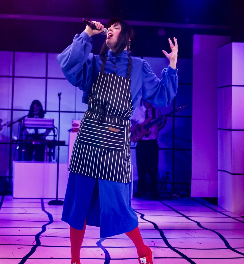 A performer wearing a blue outfit, black-and-white striped apron, red socks, and red shoes sings passionately on stage, with musicians playing in the background under purple lighting.