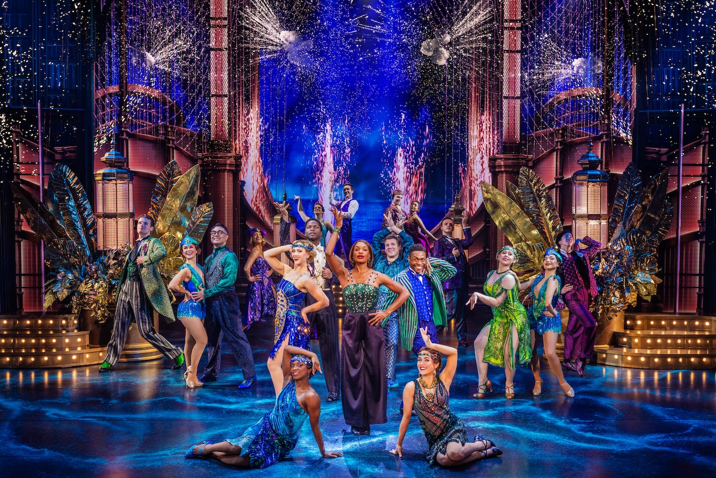 A vibrant stage performance with a diverse cast in colorful, glamorous costumes posing dramatically amid sparkling fireworks and elaborate, Art Deco-inspired set decorations.