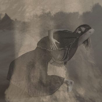 A woman in a dark, flowing dress poses dramatically with one arm behind her head and the other hand on her abdomen. The image has a grainy, sepia-toned texture, giving it a dreamy, abstract feel.