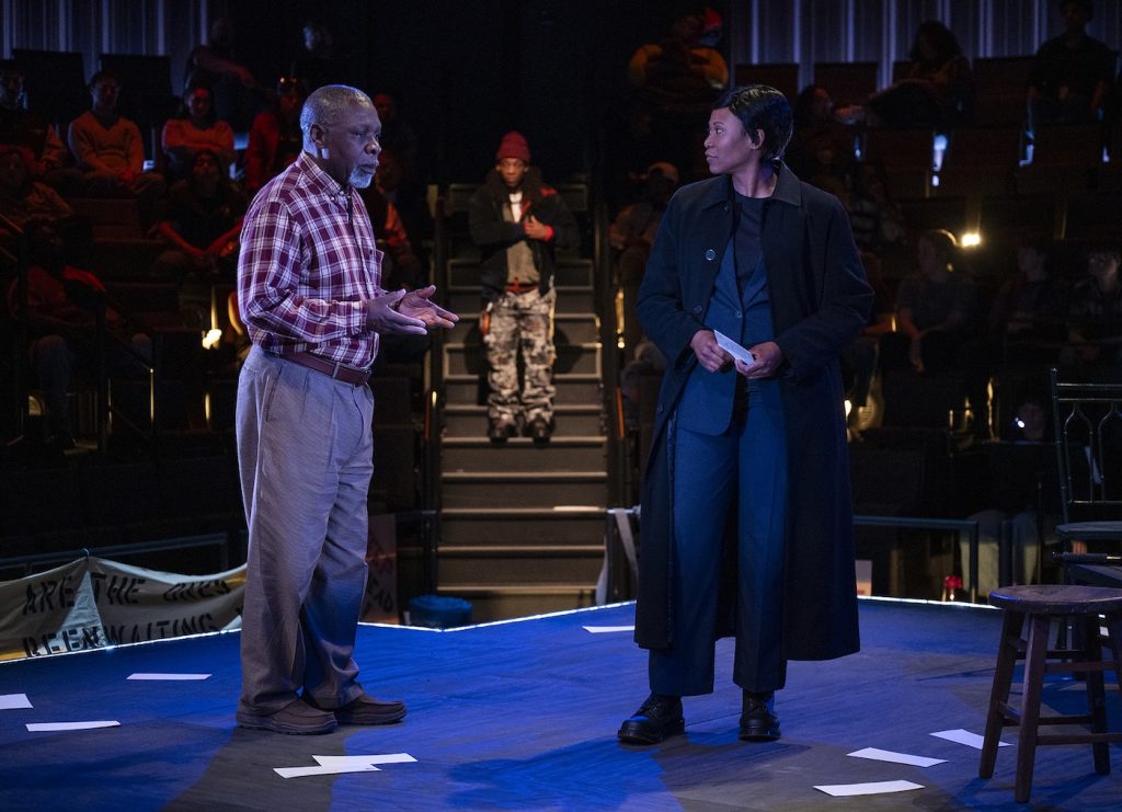 Two actors stand on a dimly lit stage facing each other, one in a red plaid shirt and khakis, the other in a long dark coat; an audience sits in the background and a person stands at the top of the stairs.