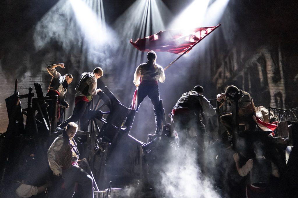 A group of people in historical clothing stand on a smoky barricade, one person raises a red flag dramatically under bright stage lights, evoking a scene of rebellion or revolution.