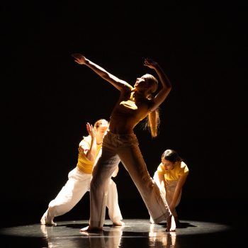 Three dancers in yellow and white outfits perform on a dark stage, dramatically lit from above. The central dancer strikes an expressive pose with arms extended, while the others pose behind and beside her.