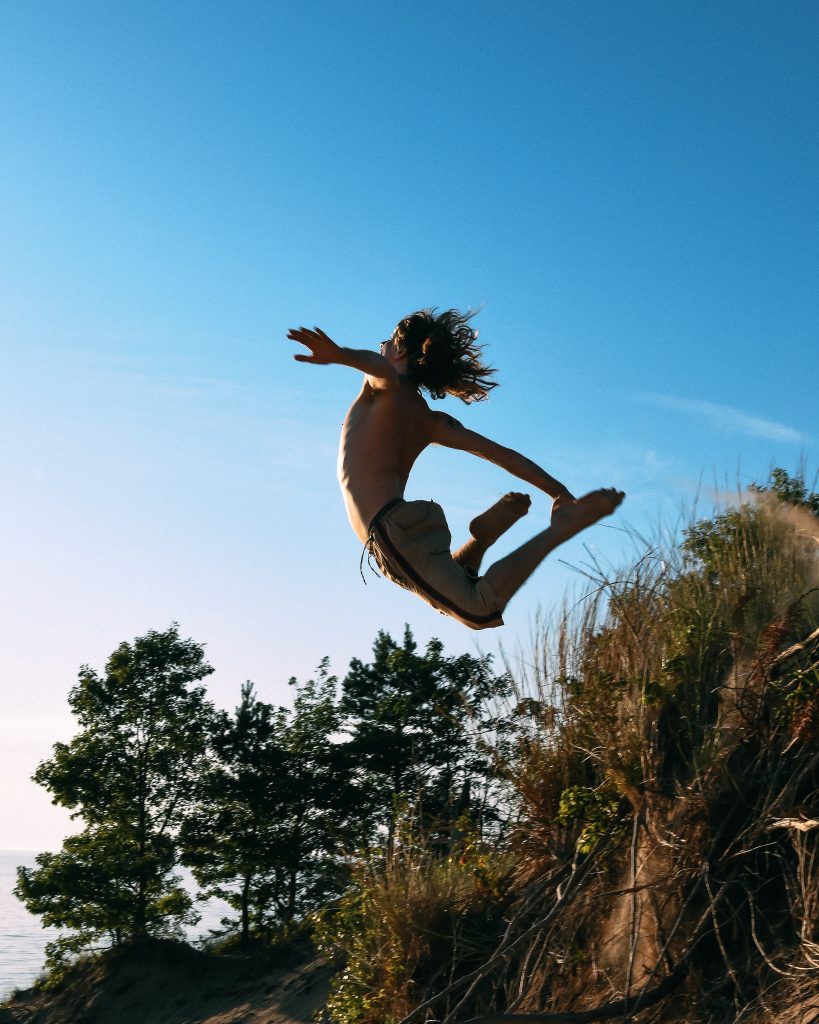 A young person with long hair, wearing shorts, leaps energetically off a sandy hill, silhouetted against a clear blue sky with trees in the background.