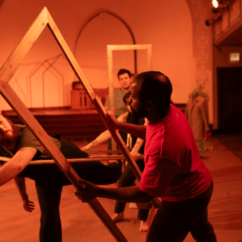Three people perform in a dimly lit room, interacting with large wooden frames. One person leans back through a frame while others hold and support it, creating a dynamic and expressive scene.