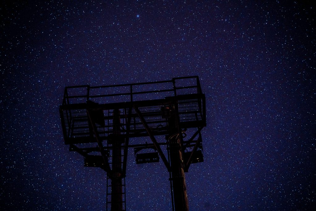 A dark silhouette of a metal structure, possibly a platform or tower, stands against a night sky filled with numerous bright stars.
