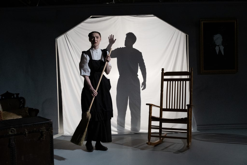 A woman in old-fashioned clothing holds a broom and stands in front of a white sheet, with a man’s shadow cast behind her. Beside her is a wooden rocking chair, a chest, and a portrait on the wall.