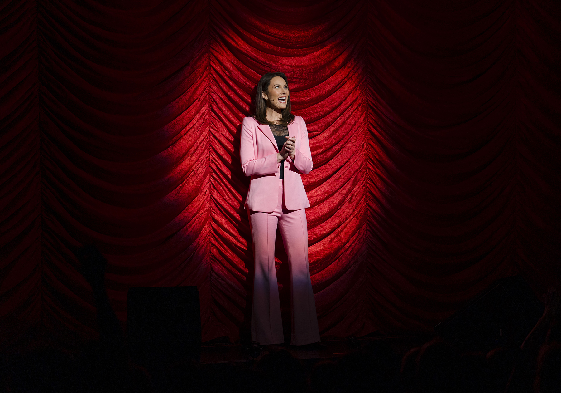 A woman in a light pink suit stands on stage holding a microphone, smiling, with dramatic red curtains and spotlight behind her.