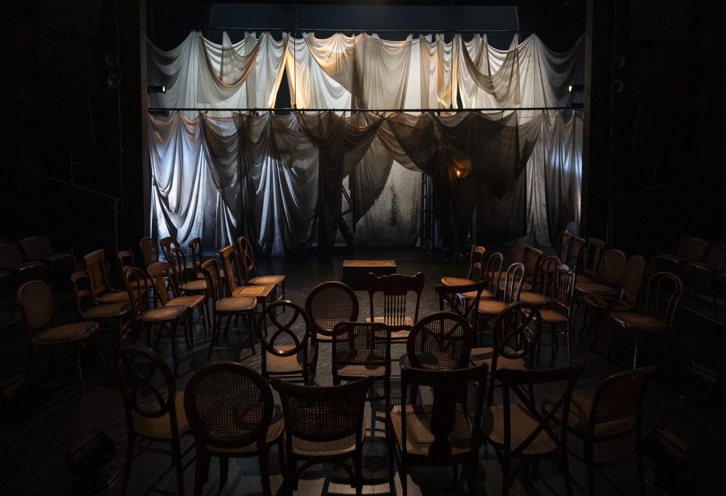 A dimly lit theater stage with sheer, draped curtains as the backdrop and rows of empty wooden chairs arranged in a semicircle facing the stage.