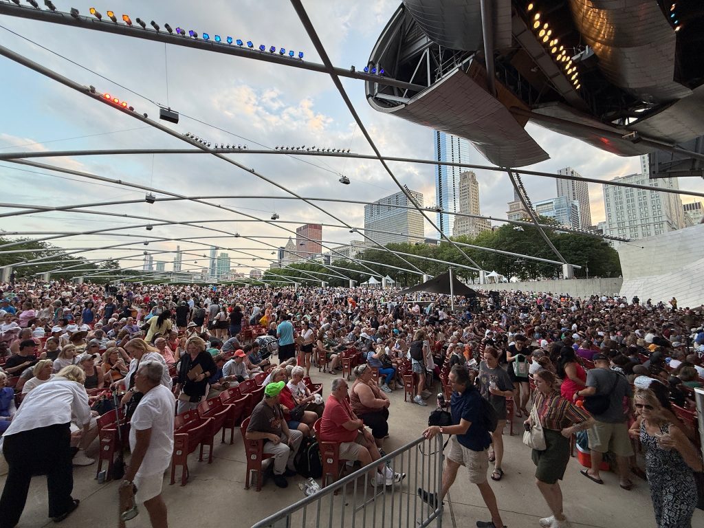 A large crowd gathers at an outdoor amphitheater in a city, seated and standing near the stage under a steel canopy, with skyscrapers and trees in the background.