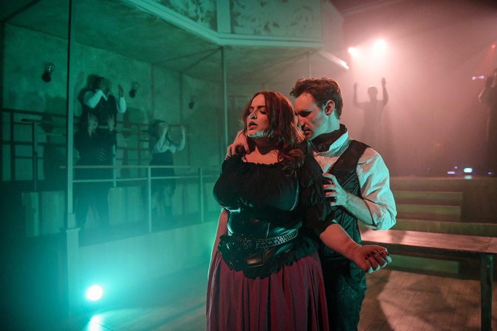 A woman in a red skirt and black top stands on stage as a man in a vest embraces her from behind. Other actors and dramatic lighting create a tense, theatrical atmosphere.