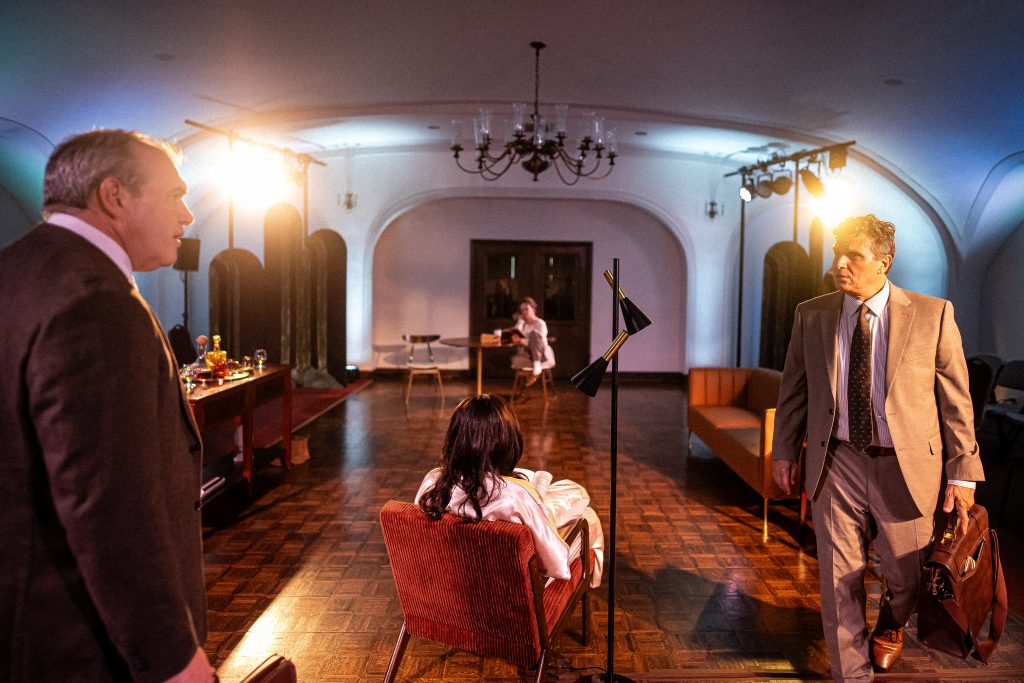 Two men in suits stand in a room with arched ceilings, facing a seated woman. Another person sits in the background at a table. The scene is dimly lit with lamps and chandeliers.