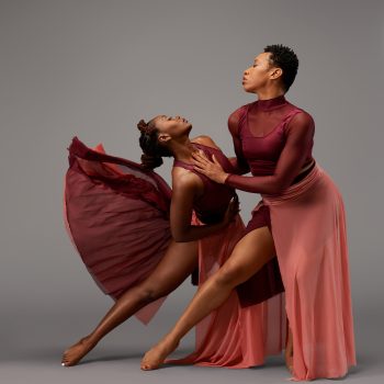 Two dancers in flowing burgundy and rose costumes pose gracefully against a gray background, one leaning back with her skirt flared, the other supporting her, both displaying expressive movement and emotion.