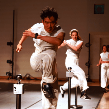 A person in white athletic clothing leaps over a small barrier indoors, followed by two others. All three appear focused and active, participating in a coordinated movement or performance.