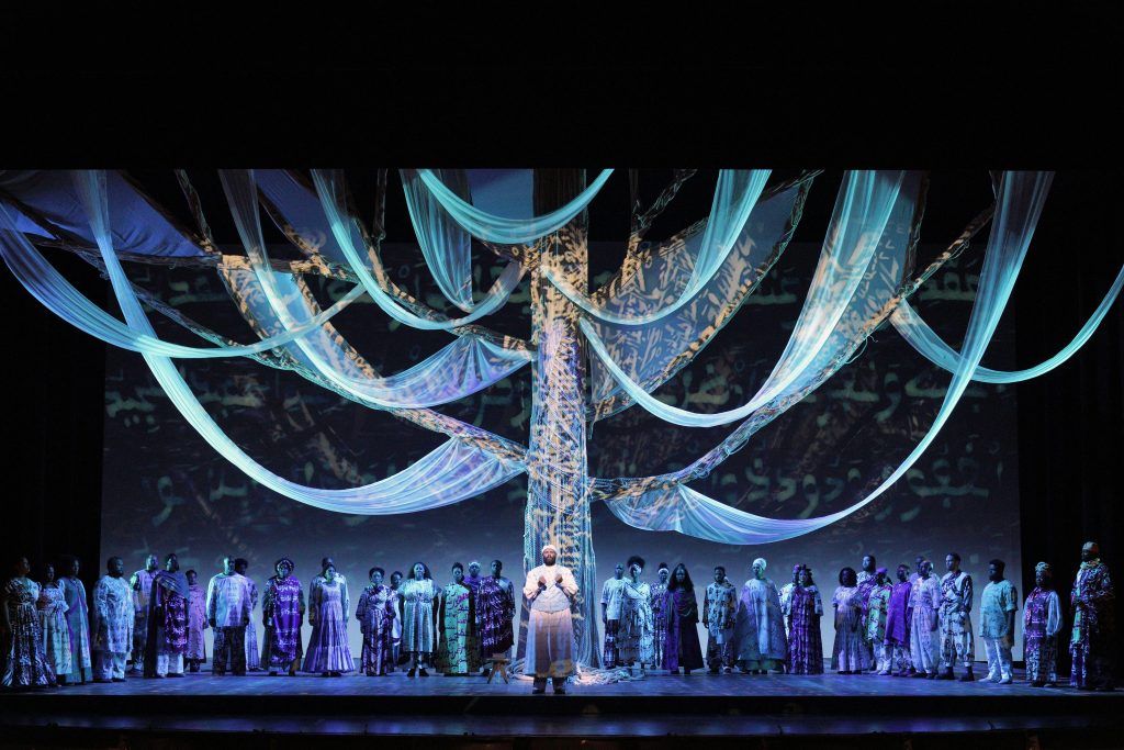 A large group of performers stands on stage around a tall, illuminated tree with flowing fabric branches, set against a blue-lit backdrop.